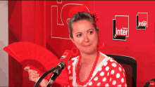 a woman in a red and white polka dot dress holds a red fan in front of a microphone with the words france inter on the wall