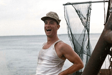 a man wearing a hat and a tank top is laughing in front of a fishing net