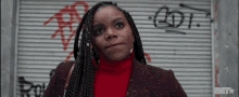 a woman with braids and a red sweater is standing in front of a garage door with graffiti on it .