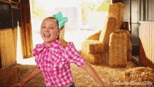 a girl in a pink and white plaid shirt with a blue bow on her head stands in front of hay bales