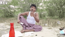 a man in a white tank top sits on the ground with his legs crossed and a red bottle of water in front of him .