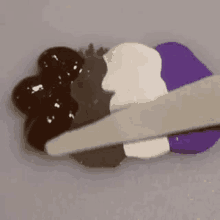 a spatula is being used to spread a purple , white , and brown paint on a piece of paper .