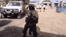 a man in a military uniform is standing in a dirt field with a white suv in the background .