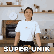 a woman in a white t-shirt stands in a kitchen with a plate of food and the words super unik above her