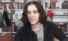 a young man with long curly hair is sitting in front of a microphone in front of a bookshelf .