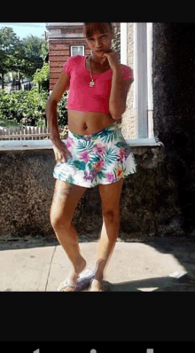 a woman in a pink crop top and floral shorts stands in front of a window