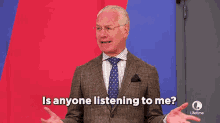 a man in a suit and tie is standing in front of a red and blue wall and asking is anyone listening to me ?