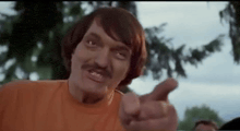 a man with a mustache and an orange shirt is pointing at the camera .