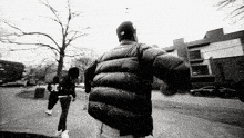 a man in a black jacket and hat is walking down a sidewalk in a black and white photo .