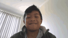 a young boy in a green jacket smiles in front of a window with vertical blinds