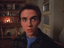 a young man in a plaid shirt and blue shirt looks at the camera