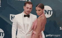 a man in a white suit and bow tie stands next to a woman in a pink dress on a red carpet that says nt