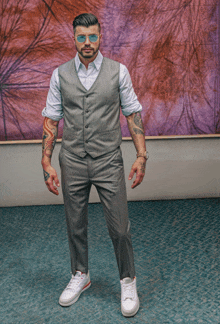 a man wearing a suit vest and white sneakers stands in front of a purple wall