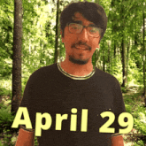 a man wearing glasses and a black shirt stands in the woods on april 29
