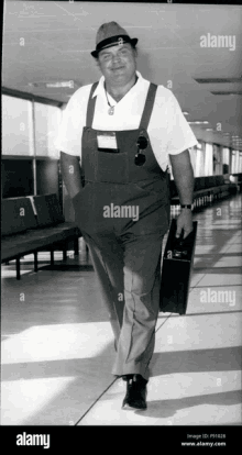 a man wearing overalls and a hat is walking down a hallway holding a suitcase