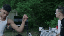 a man in a white tank top is flexing his muscles in front of a group of people at a picnic table .