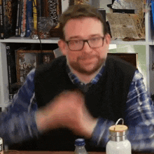 a man wearing glasses and a plaid shirt is clapping his hands in front of a bookshelf with a book titled fantasy roleplay