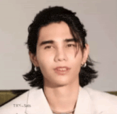 a young man with long hair and earrings is wearing a white shirt and looking at the camera .