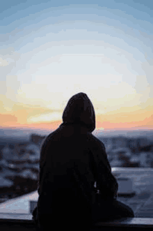 a person in a hoodie is sitting on a balcony looking at the sunset .