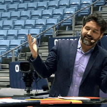 a man in a suit and a microphone stands in front of an empty stadium with a sign that says o on it