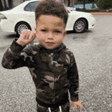 a young boy in a camouflage jacket is standing in front of a white car and giving the middle finger .