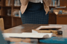 a woman in a plaid skirt is standing in front of a table with books on it in a library .