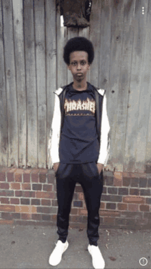 a man wearing a thrasher shirt stands in front of a wooden fence