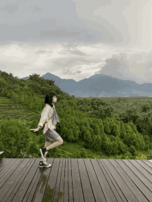 a woman wearing a mask stands on a wooden deck overlooking a lush green forest