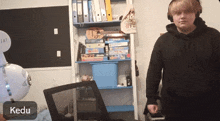 a man wearing headphones stands in front of a shelf full of binders and a chair with the name kedu written on it