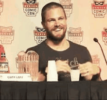 a man with a beard is sitting at a table in front of a microphone holding cups .