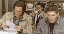 two men are sitting at a table looking at a piece of paper and one is taking a picture of the other .