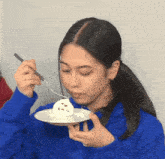 a woman in a blue sweatshirt is holding a plate of food with a fork in her mouth