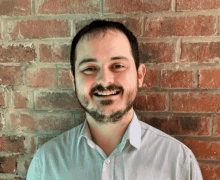 a man with a beard is smiling in front of a red brick wall