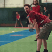 a man in a red shirt stands on a basketball court with la guarimba film festival written on the bottom left