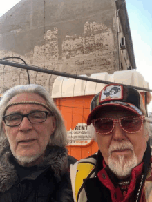 two men are posing for a picture in front of an orange sign that says rent