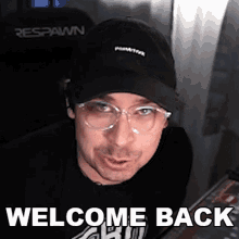 a man wearing glasses and a baseball cap is standing in front of a sign that says `` welcome back '' .