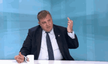 a man in a suit and tie is sitting at a table with a cup of coffee and pointing at something .