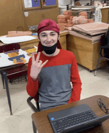 a young man wearing a red headband is sitting at a desk with a laptop and giving a peace sign