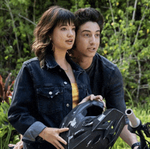 a woman in a denim jacket holds a helmet next to a man on a bike