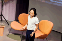 a man in a white shirt stands in front of an orange chair with visitors can experience written on the screen behind him