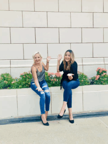 two women sitting next to each other in front of a wall with flowers