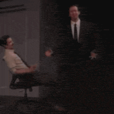 a man in a suit and tie is standing next to another man in a chair .
