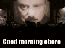 a man with a mustache is standing in front of a large screen with the words `` good morning oboro '' .