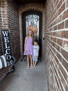 two children standing in front of a brick house with a welcome sign