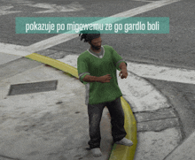 a man in a green shirt is standing on a sidewalk with the words pokazuje po migowemu ze go gardlo boli written above him