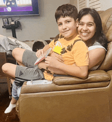 a woman sits on a couch holding a young boy who is wearing a yellow shirt with a crown on the back