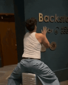 a man in a white tank top and blue jeans is kneeling down in front of a wall with the word back on it .