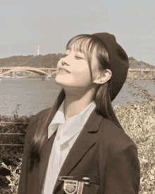 a young woman wearing a suit and a beret is standing in front of a body of water .