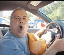 a man in a blue shirt is driving a car and holding a bag of food in his mouth