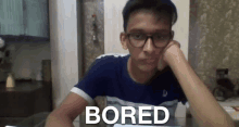 a young man wearing glasses is sitting at a table with his hand on his chin and the word bored written on the screen .